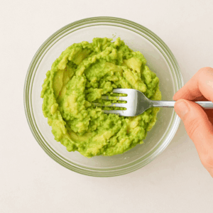 Mão amassando abacate maduro com garfo em tigela de vidro para preparar guacamole