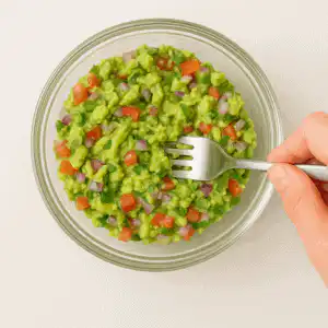 Mão misturando abacate com tomate, cebola e coentro em tigela de vidro com garfo